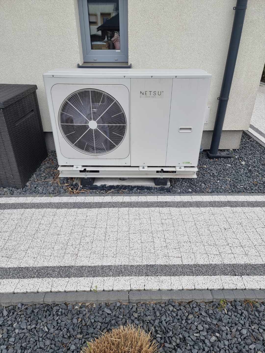 Biała pompa ciepła marki Netsu zamontowana na zewnątrz budynku, widoczna na tle elewacji z oknem i szarego chodnika z kamykami.