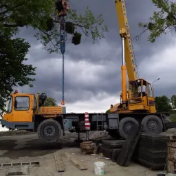 Żółty dźwig samochodowy z wysięgnikiem podnosi ciężki ładunek łańcuchami na tle pochmurnego nieba i drzew.