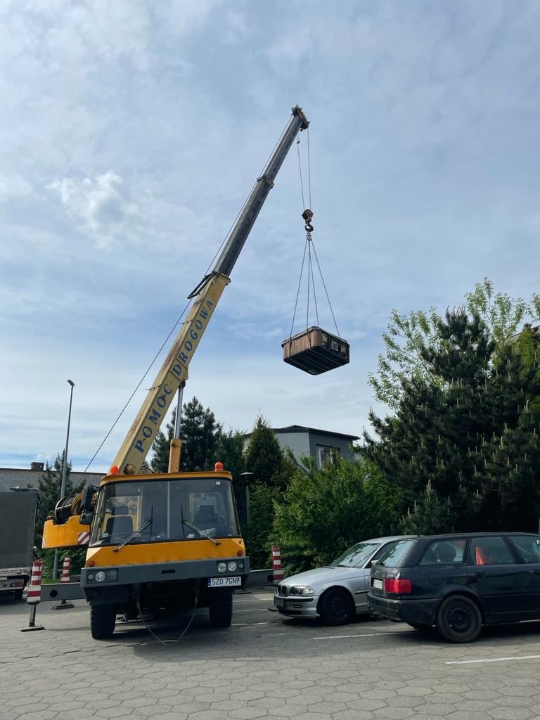 Żółty dźwig samochodowy z napisem 'POMOC DROGOWA' podnosi drewnianą kabinę w otoczeniu zaparkowanych samochodów i zieleni.