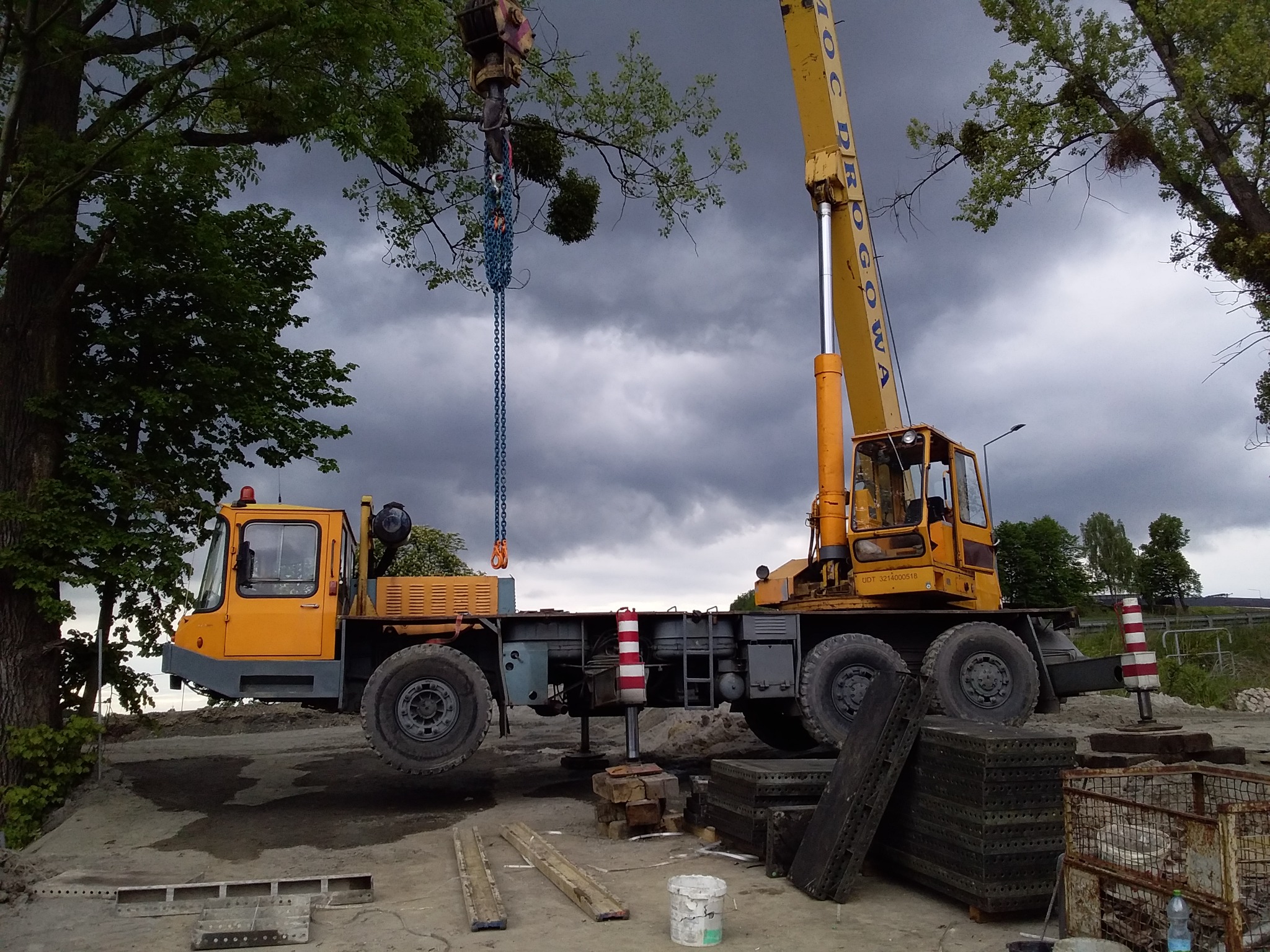 Żółty dźwig samochodowy z wysięgnikiem podnosi ciężki ładunek łańcuchami na tle pochmurnego nieba i drzew.