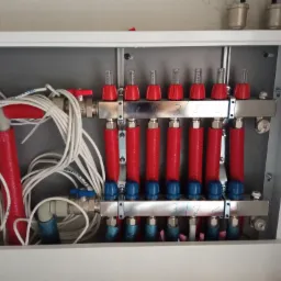 Rozdzielacz ogrzewania podłogowego w białej szafce, z czerwonymi rurami i niebieskimi zaworami, widoczne białe przewody i elementy instalacji.