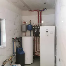 Nowoczesna kotłownia z białą pompą ciepła Panasonic, zbiornikiem buforowym i zmiękczaczem wody, widoczne rury miedziane i instalacja wodna na tle marmurowych płytek w surowym wnętrzu.