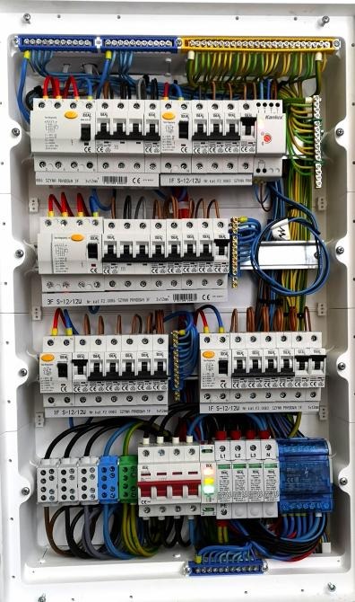 Otwarta skrzynka rozdzielcza z bezpiecznikami, wyłącznikami różnicowoprądowymi i listwami zaciskowymi, uporządkowane kable w kolorach niebieskim, żółto-zielonym i brązowym, widoczne oznaczenia...