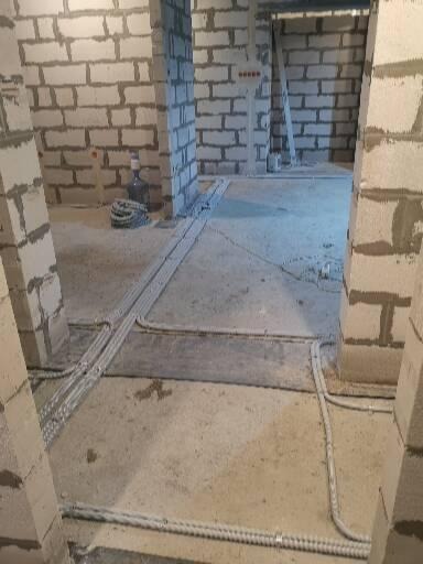 Surowy stan wnętrza w trakcie układania przewodów elektrycznych pod tynkiem, widoczne szare ściany z betonu komórkowego i kable w peszlach na podłodze.