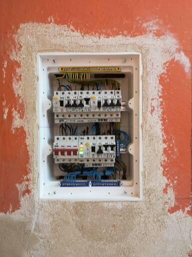 Wewnętrzna skrzynka bezpiecznikowa z zainstalowanymi wyłącznikami, zamontowana w pomarańczowej ścianie z widocznymi śladami po szpachlowaniu.