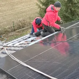 Montaż paneli słonecznych na dachu z dachówki ceramicznej; dwóch instalatorów w czerwonych kurtkach i czapkach roboczych mocuje panele do aluminiowych szyn montażowych.