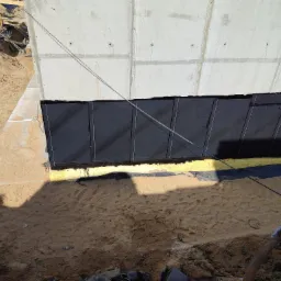 Betonowy fundament z nałożoną czarną izolacją przeciwwilgociową i żółtą warstwą ochronną, widok z góry na wykop z piaskiem.