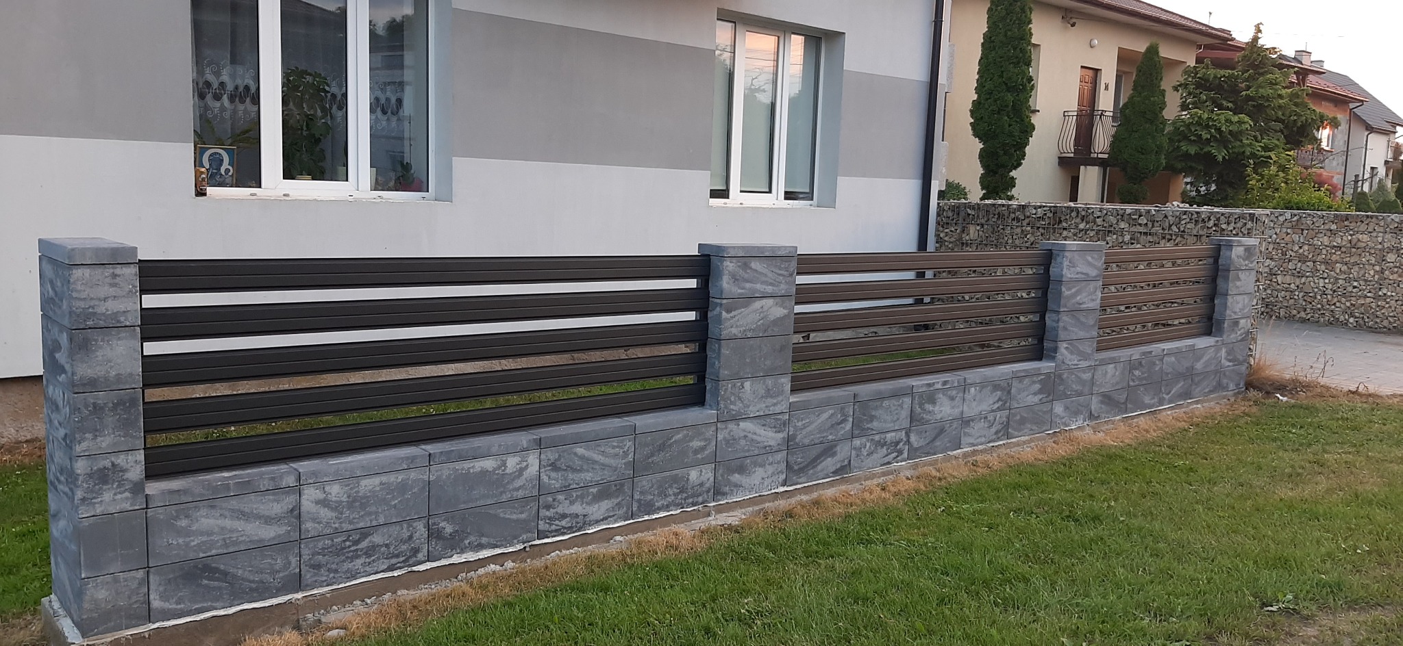 Nowoczesne ogrodzenie panelowe z poziomymi przęsłami w kolorze grafitowym, osadzone na podmurówce z szarych bloczków betonowych, na tle domu z białymi oknami i zielenią trawnika.