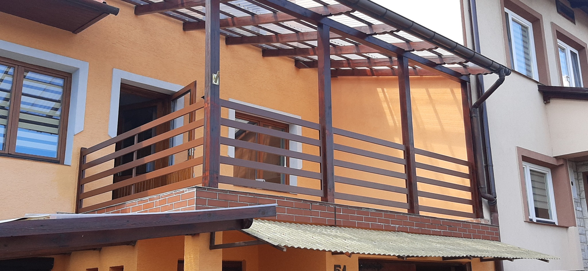 Zadaszony balkon z drewnianą balustradą i częściowo odsłoniętym oknem, widok z dołu na budynek z pomarańczową elewacją i oknami z żaluzjami.