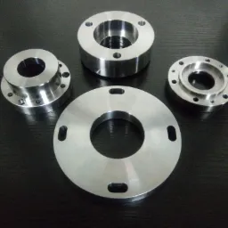 Cztery precyzyjnie obrobione, metalowe elementy kołnierzowe z otworami, ułożone na ciemnym, lekko połyskującym blacie, ukazujące detale obróbki CNC.