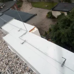 Widok z góry na płaski dach pokryty białym materiałem izolacyjnym i obciążony kamieniami, widoczne metalowe elementy mocujące i fragmenty innych budynków w tle.
