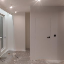 Kobasvar bud - Wnętrze po świeżym pomalowaniu na biało, widoczne ślady farby na podłodze, puszka z farbą i wałek malarski w rogu pokoju, biały grzejnik pod oknem, minimalistyczne drzwi wewnętrzne.