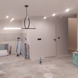 Kobasvar bud - Wnętrze w trakcie prac wykończeniowych: świeżo pomalowane białe ściany, nowoczesna lampa sufitowa, przygotowane panele podłogowe Quick Step, widoczne ślady gładzi na podłodze, butelka wody...