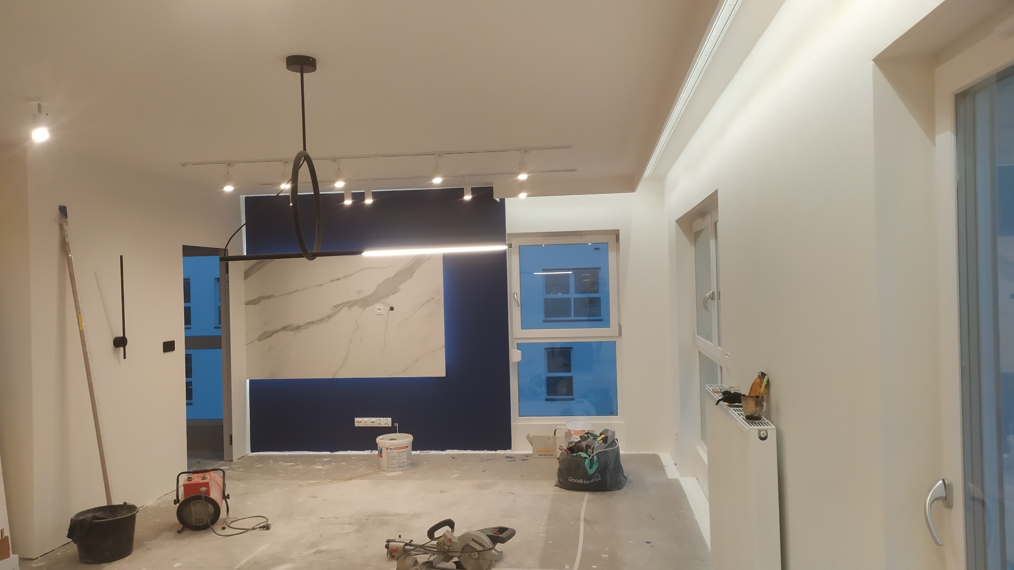 Wnętrze w trakcie prac wykończeniowych: świeżo pomalowane białe ściany, niebieska ściana z marmurową płytą, nowoczesne oświetlenie sufitowe, widoczne narzędzia i akcesoria malarskie na podłodze...
