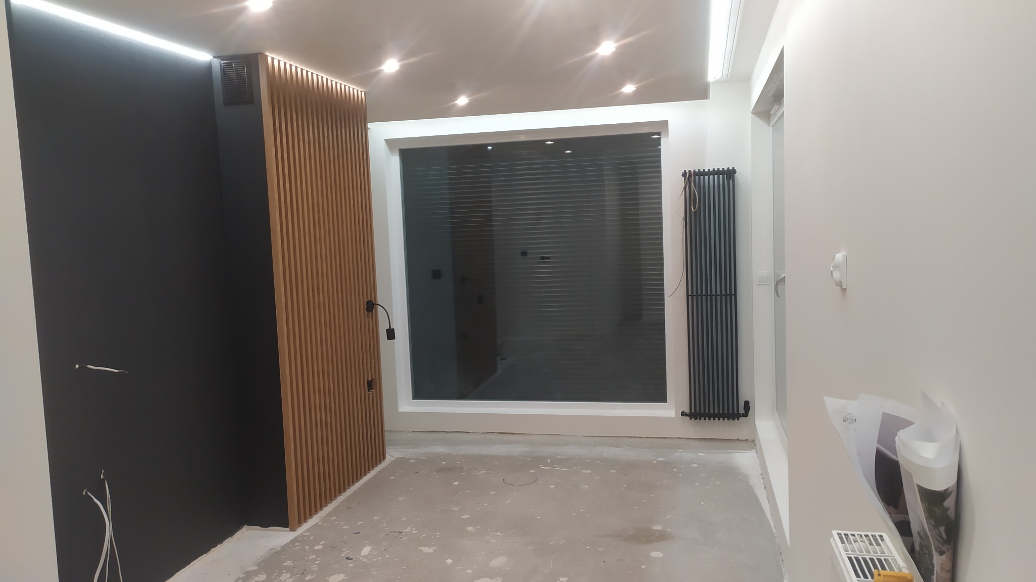 Wnętrze w trakcie prac wykończeniowych: czarna ściana z drewnianymi lamelami, duże okno z żaluzjami, grzejnik panelowy i rozłożone plany remontowe.