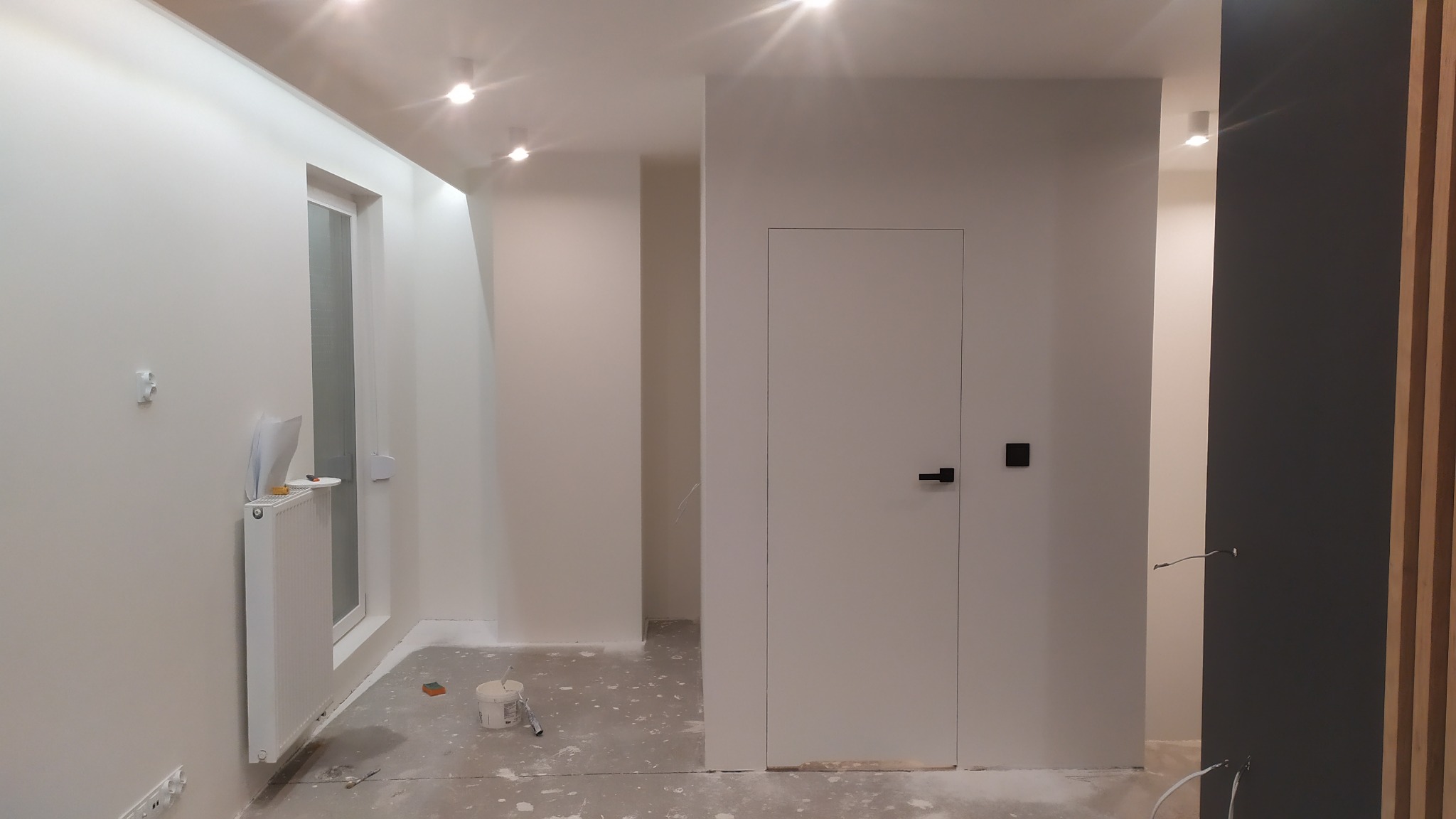 Wnętrze po świeżym pomalowaniu na biało, widoczne ślady farby na podłodze, puszka z farbą i wałek malarski w rogu pokoju, biały grzejnik pod oknem, minimalistyczne drzwi wewnętrzne.