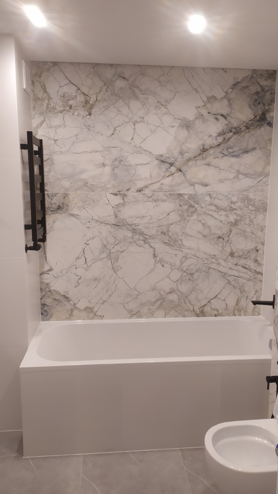 Wykończona łazienka z wanną obudowaną panelami, ścianą wyłożoną płytkami imitującymi marmur oraz czarnym grzejnikiem drabinkowym.