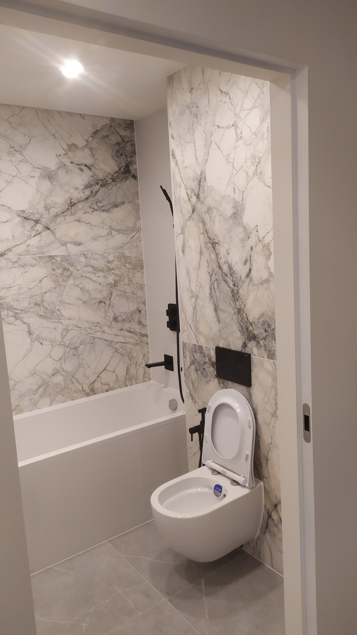 Wykończona łazienka we Wrocławiu z wanną i podwieszaną toaletą, wykończona płytkami imitującymi marmur z czarną armaturą, oświetlona punktowo.