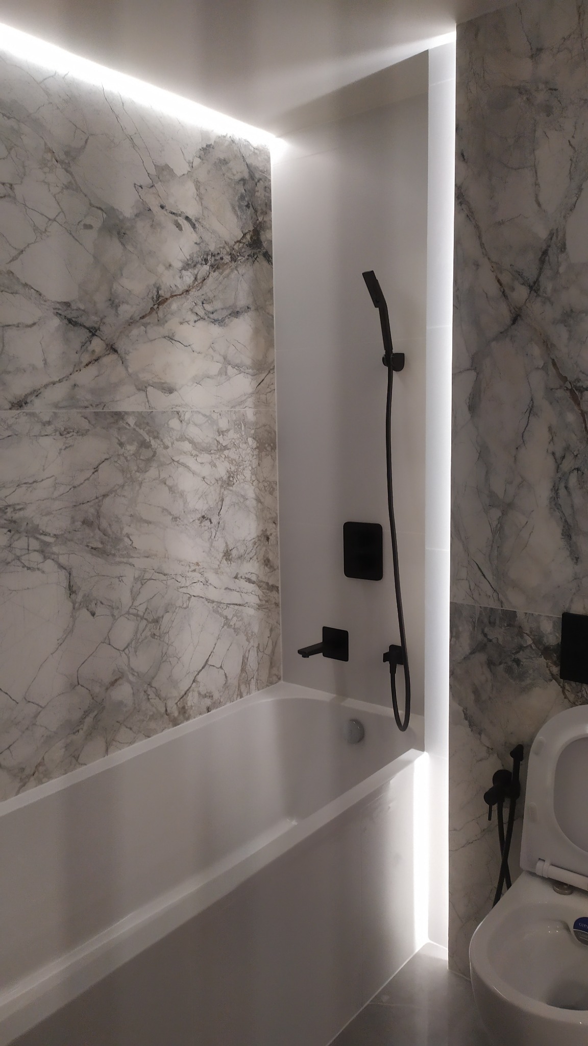 Nowoczesna łazienka z wanną, marmurowymi płytkami i czarną armaturą, oświetlenie LED w pionowej wnęce.