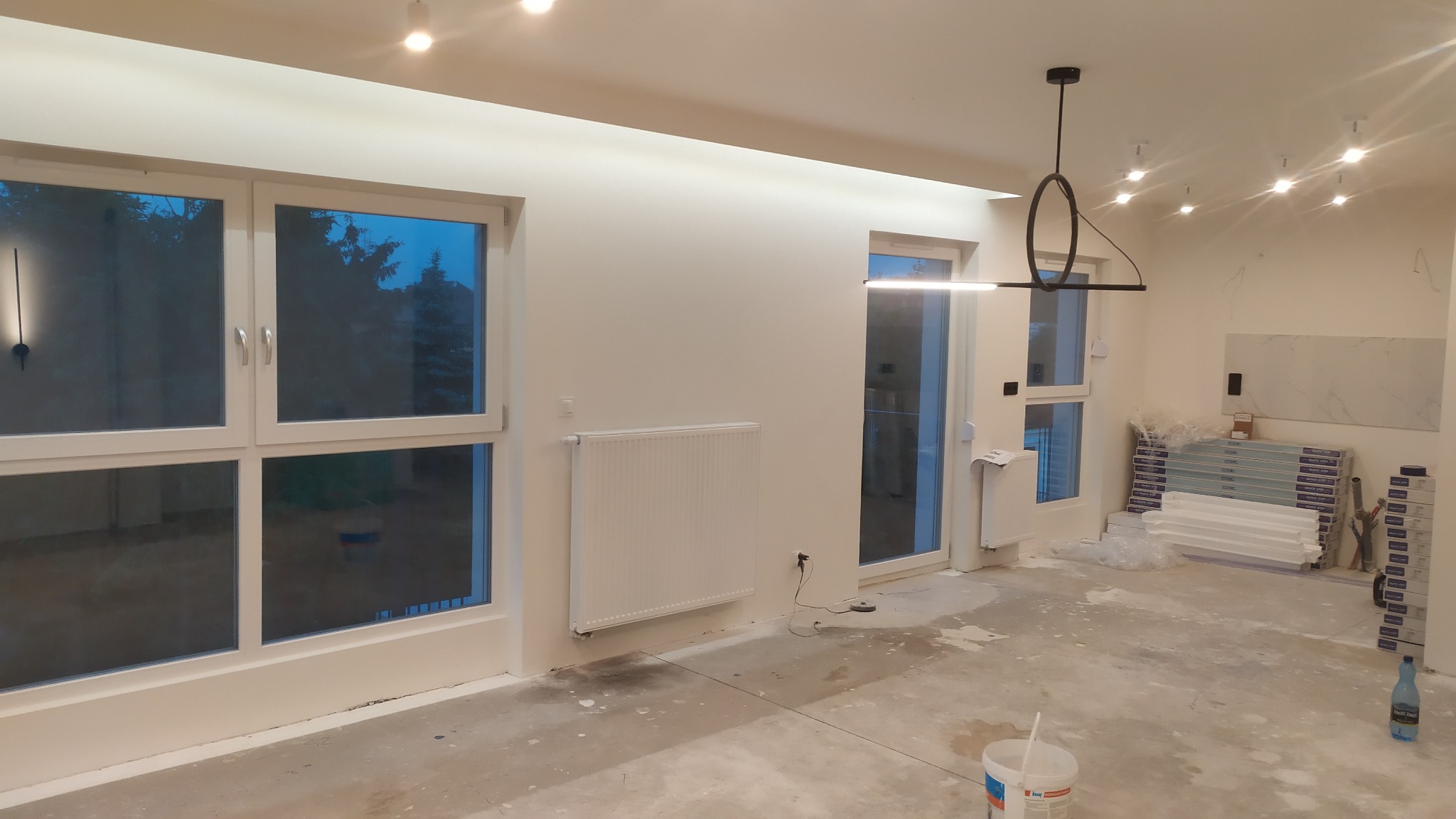 Wnętrze w trakcie prac wykończeniowych: świeżo pomalowane ściany, grzejnik, okna, nowoczesna lampa, podłoga przygotowana do dalszych prac, widoczne materiały budowlane.