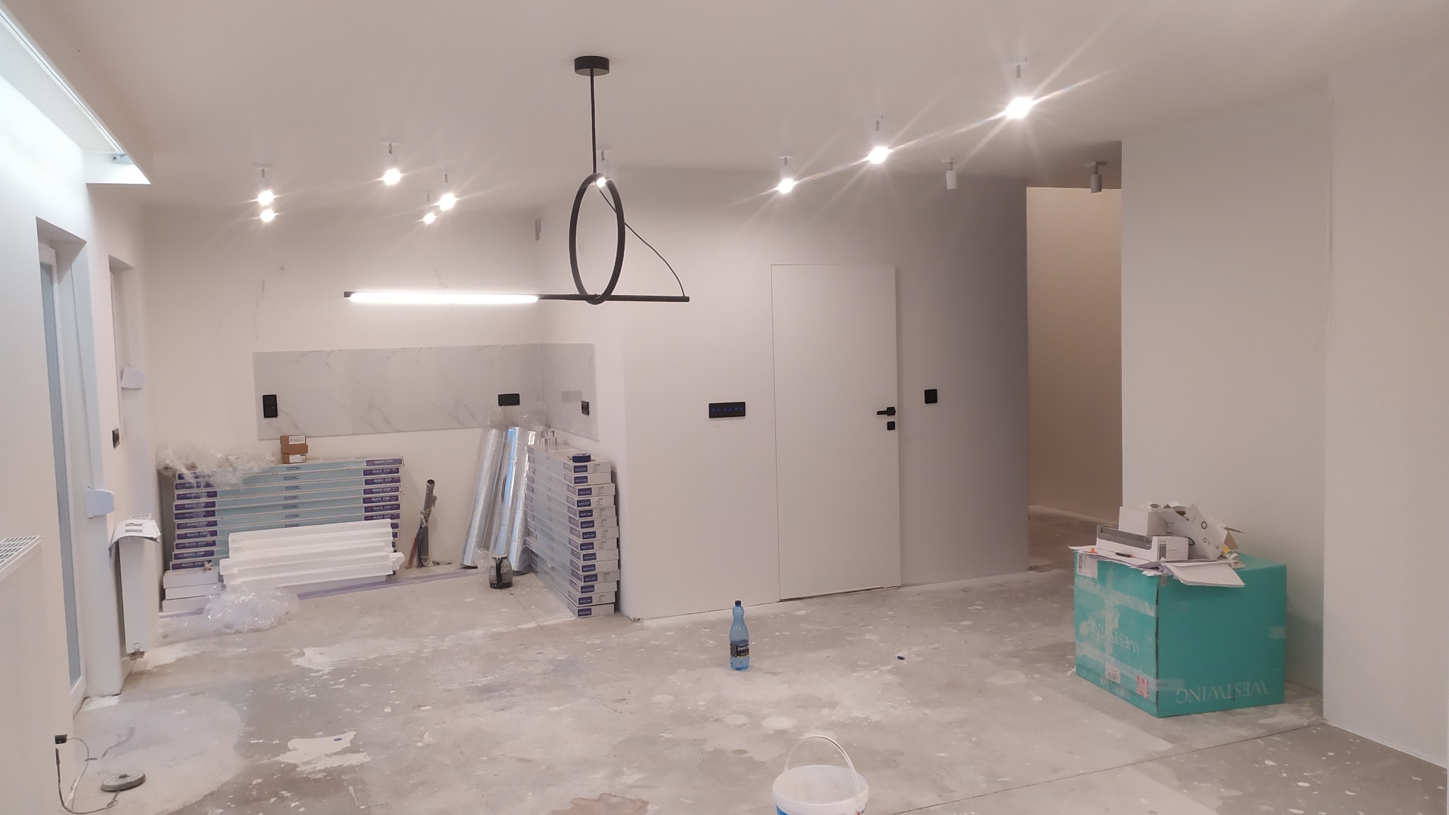 Wnętrze w trakcie prac wykończeniowych: świeżo pomalowane białe ściany, nowoczesna lampa sufitowa, przygotowane panele podłogowe Quick Step, widoczne ślady gładzi na podłodze, butelka wody...