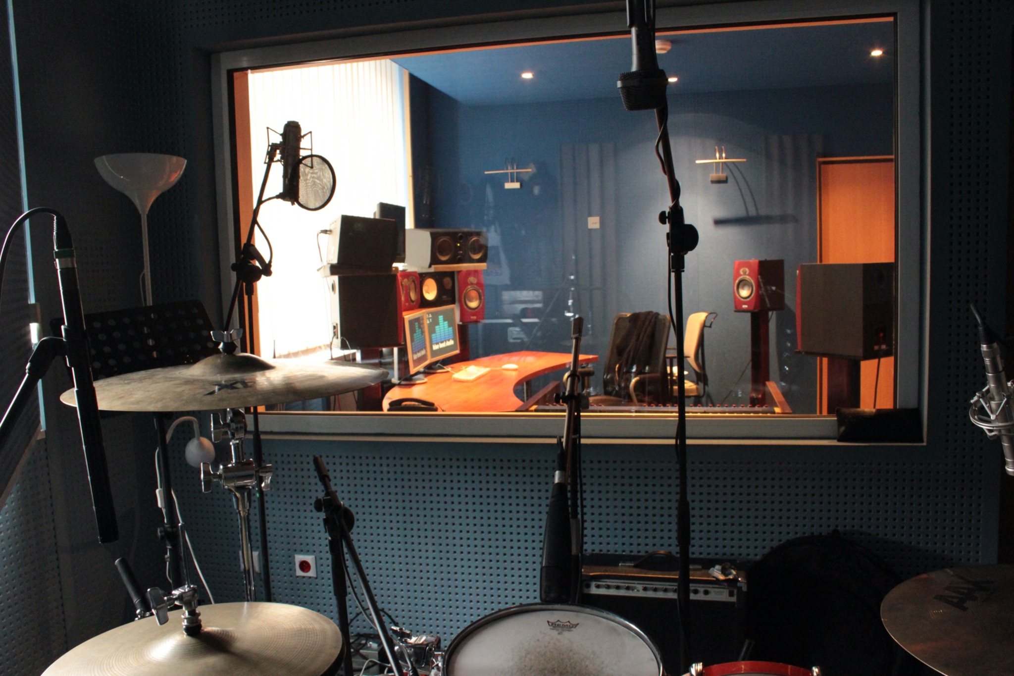 Wnętrze studia nagrań z widocznym zestawem perkusyjnym na pierwszym planie oraz reżyserką za szybą, z monitorami, głośnikami i biurkiem w kolorze drewna.