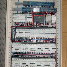Rozdzielnica elektryczna z licznymi bezpiecznikami, przewodami w kolorach niebieskim, czerwonym i czarnym, oraz modułem ABB OT63F3C, zamontowana w białej obudowie.