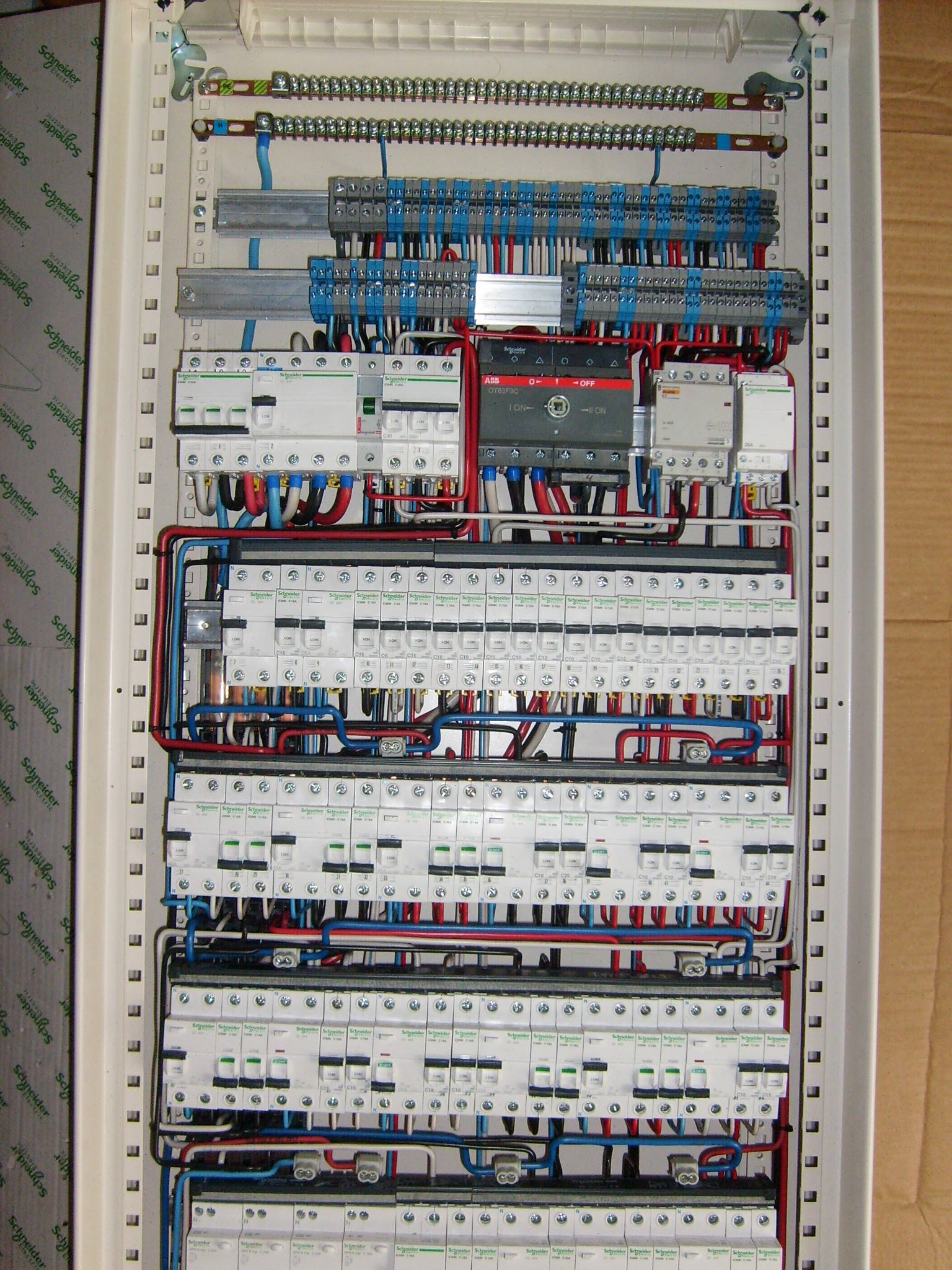 Rozdzielnica elektryczna z licznymi bezpiecznikami, przewodami w kolorach niebieskim, czerwonym i czarnym, oraz modułem ABB OT63F3C, zamontowana w białej obudowie.