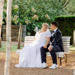 Para młoda siedzi na ławce w parku, panna młoda w białej sukni i sportowych butach, pan młody w granatowym garniturze i muszce, patrzą na siebie z uśmiechem, w tle zielona roślinność i brzozowe...