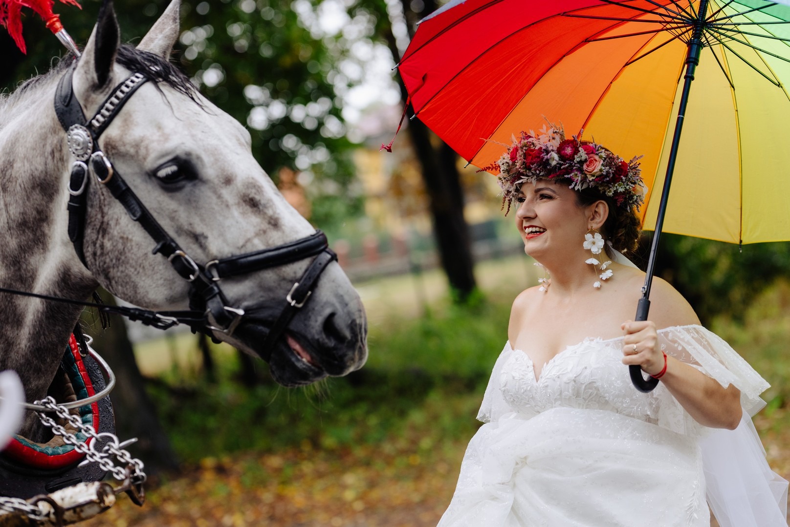 Uśmiechnięta panna młoda z wiankiem na głowie i kolorowym parasolem, obok głowa konia w uprzęży, sesja ślubna w Byczynie.