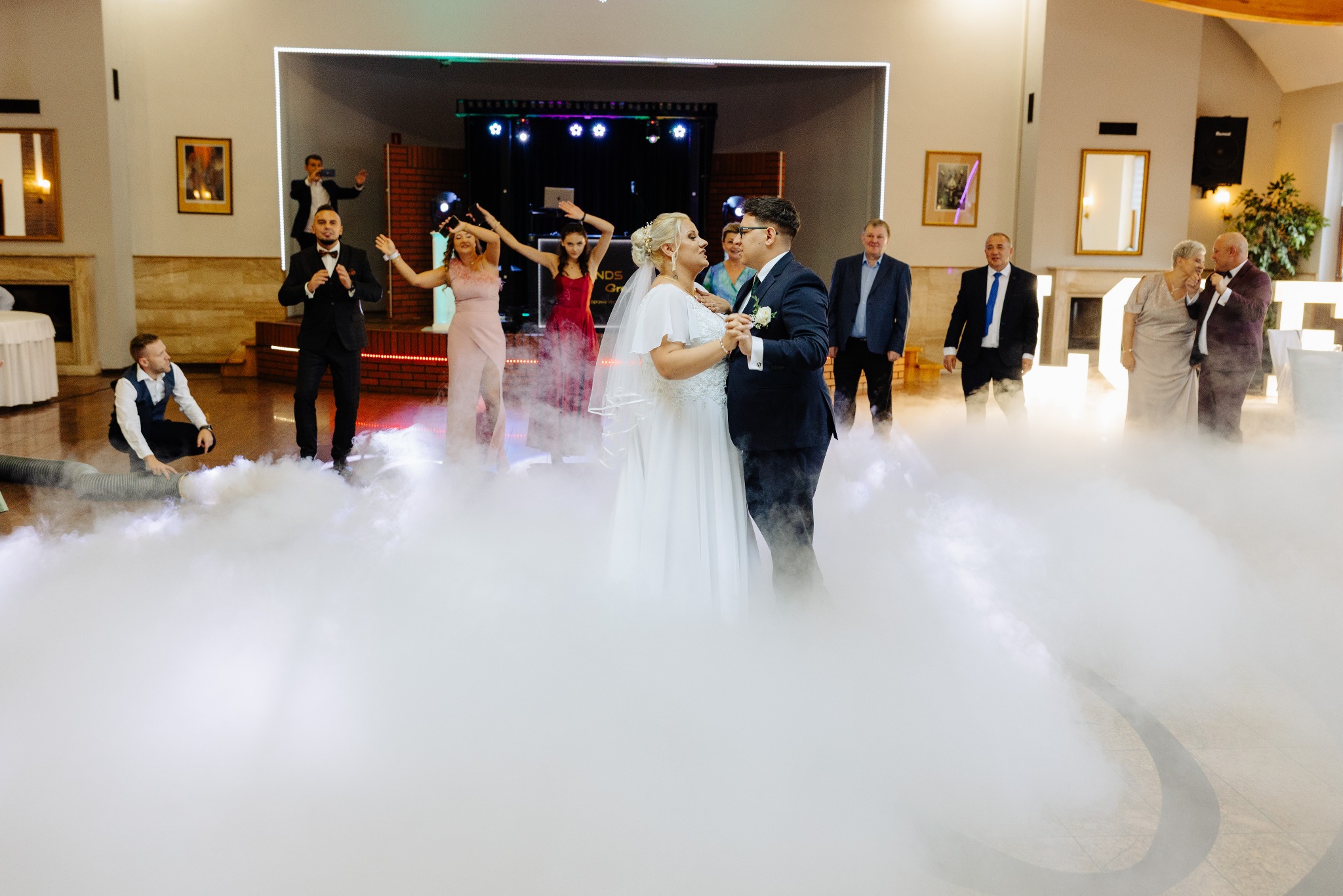 Para młoda w pierwszym tańcu, otoczona sztuczną mgłą, w tle goście weselni i oświetlenie sceniczne.