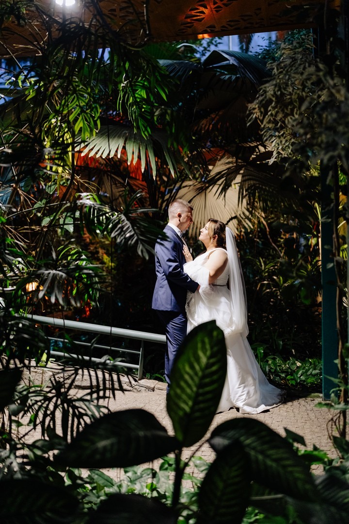 Para młoda w objęciach, otoczona bujną zielenią tropikalnej szklarni, ciepłe światło podkreśla intymną atmosferę.