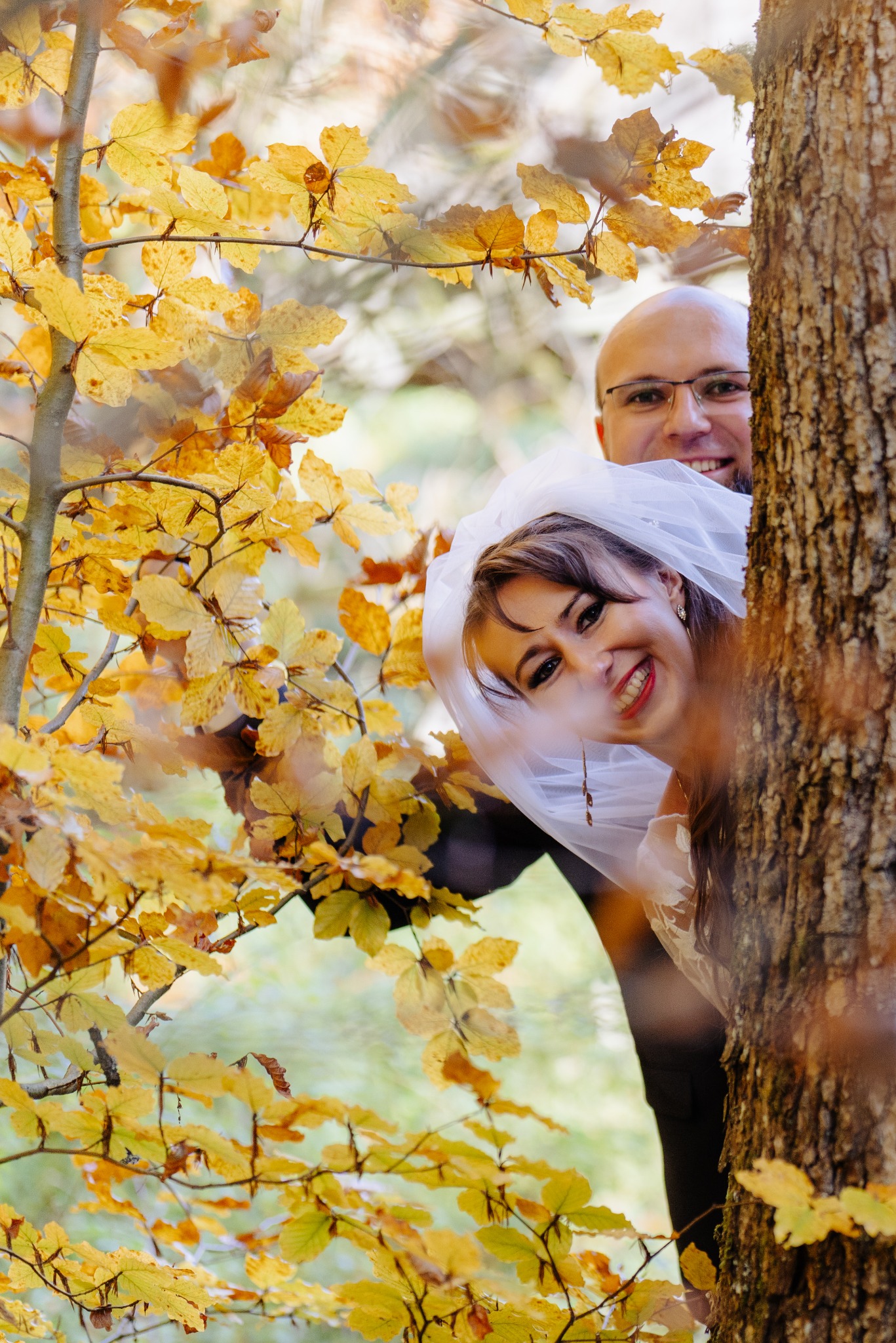 Uśmiechnięta para młoda, panna młoda w welonie i pan młody w okularach, wyglądają zza drzewa w jesiennej scenerii z żółtymi liśćmi.