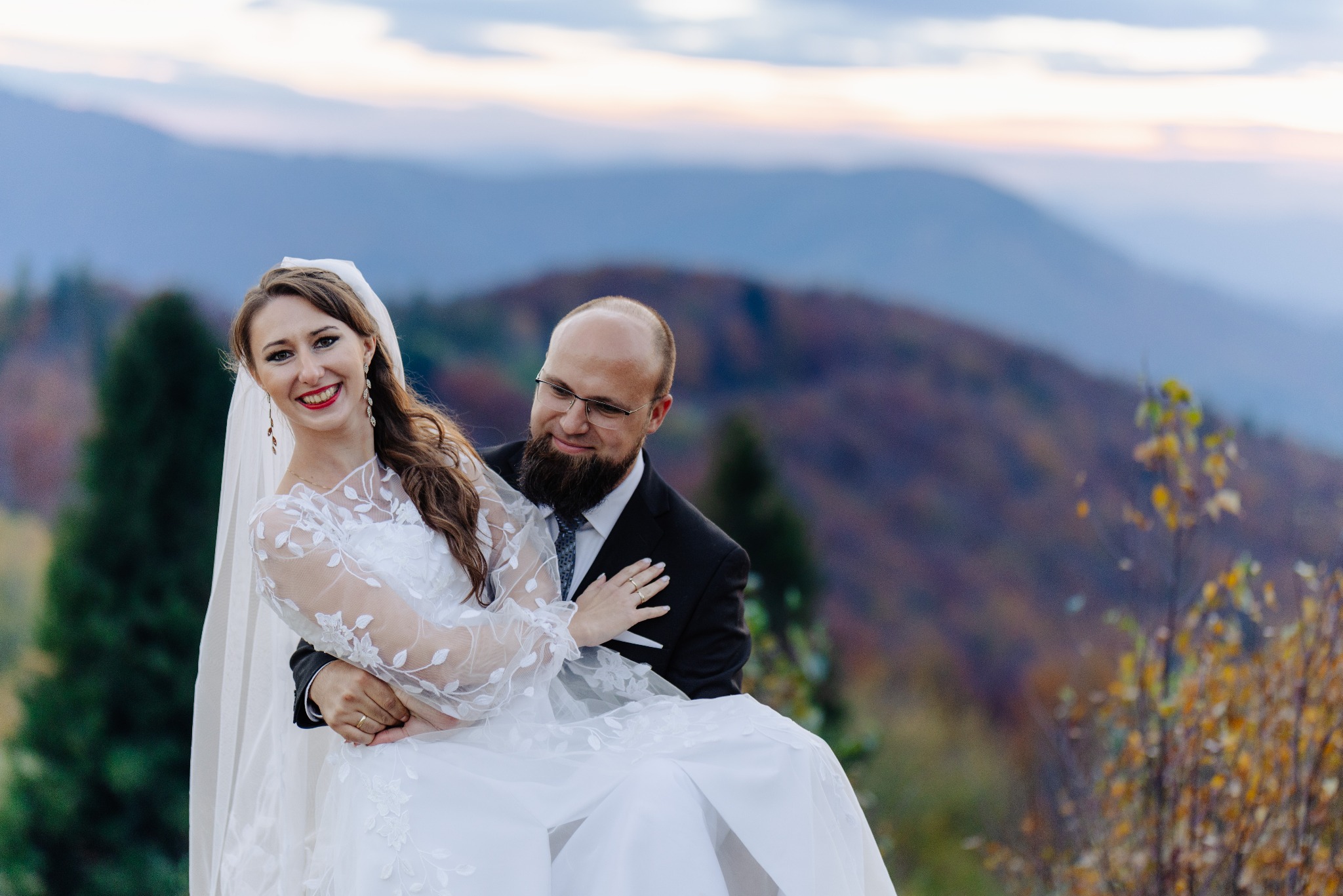 Uśmiechnięta panna młoda w białej sukni z koronkowymi rękawami, niesiona przez pana młodego na tle górskiego krajobrazu o zmierzchu.
