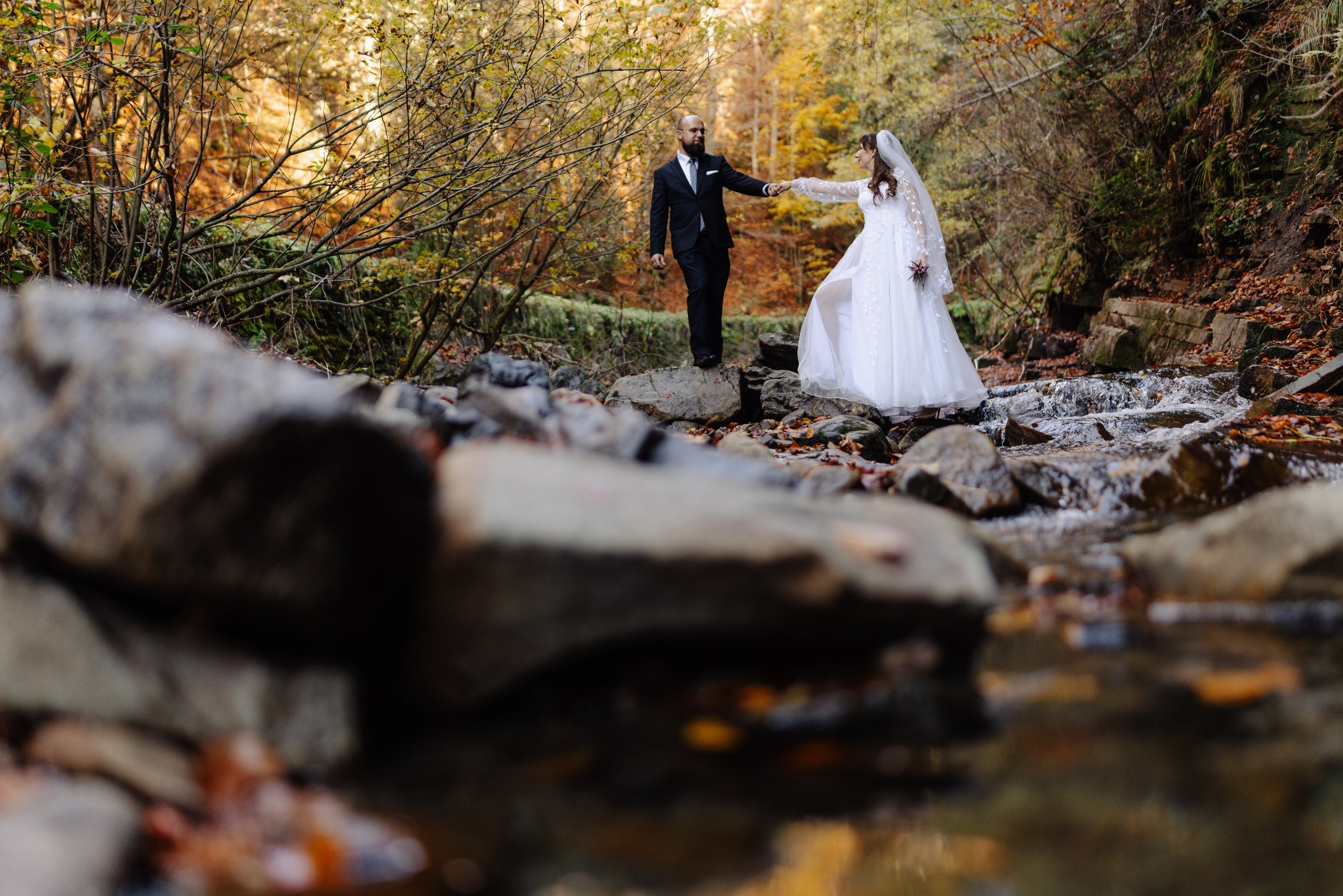 Para młoda w strojach ślubnych pozuje na kamieniach w potoku, w tle jesienny las z żółtymi liśćmi.