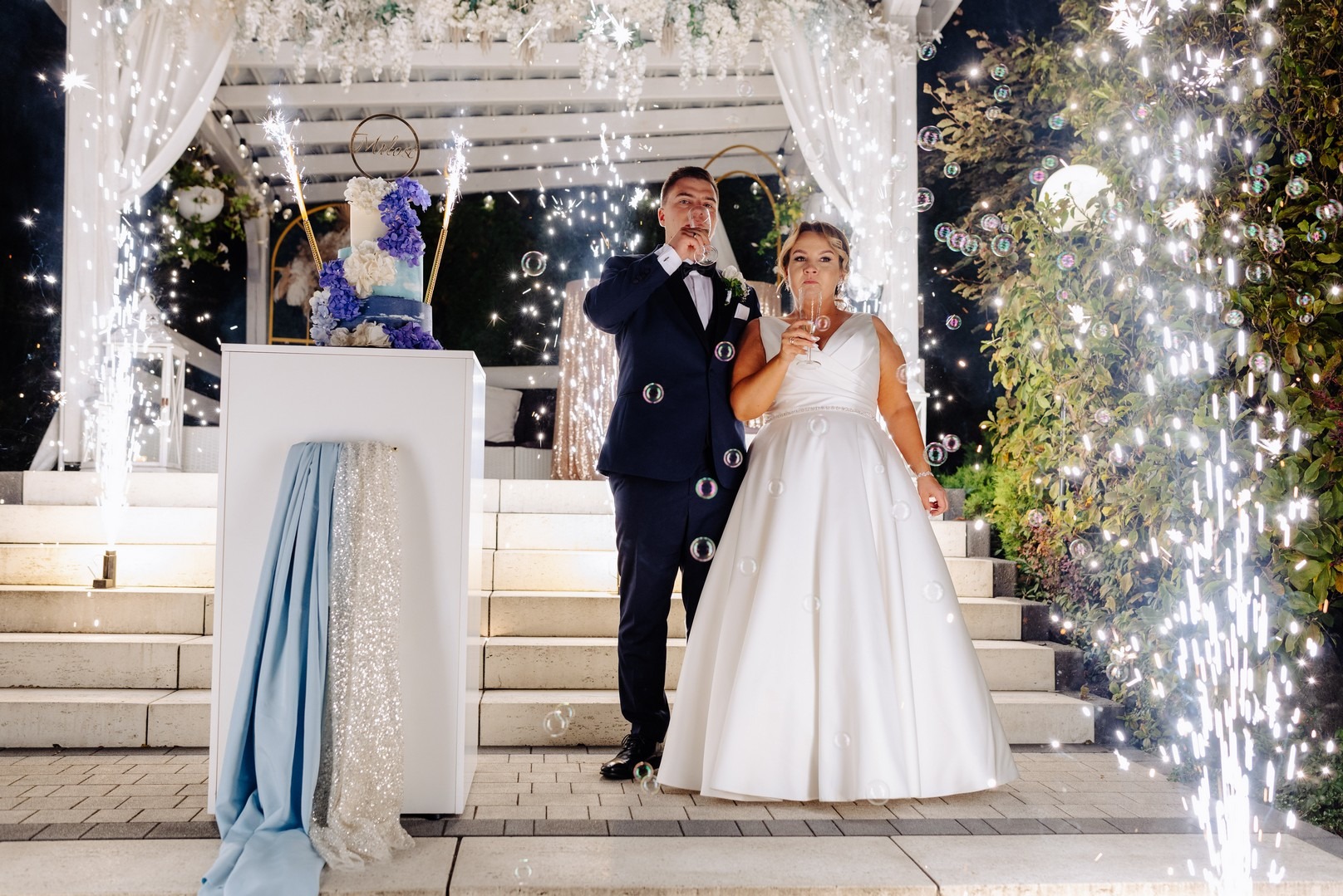 Para młoda w strojach ślubnych wznosi toast szampanem na tle tortu, fontann iskier i baniek mydlanych.