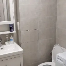 Wykończona łazienka w szarościach z umywalką w szafce, toaletą i pralką; widoczna kratka wentylacyjna na ścianie.