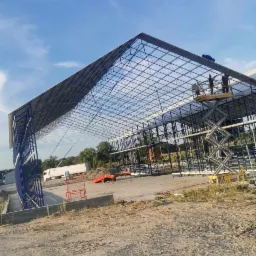 Montaż konstrukcji stalowej hali z wykorzystaniem podnośnika koszowego i płyt warstwowych na dachu, widoczne elementy konstrukcyjne i pracownicy na platformie.