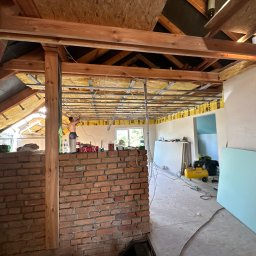 KRISPROF - Wnętrze poddasza w trakcie adaptacji: widoczne ocieplenie wełną mineralną, metalowa konstrukcja pod płyty gipsowo-kartonowe, fragment ściany z cegły, kompresor i płyta kartonowo-gipsowa oparta...