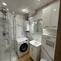 Ukończona łazienka z kabiną prysznicową, pralką Bosch, umywalką z szafką, toaletą podwieszaną oraz białymi szafkami wiszącymi, wykończona płytkami imitującymi marmur i drewnopodobną podłogą,...