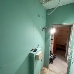 Ściana z zielonych płyt kartonowo-gipsowych w trakcie montażu, widoczne instalacje hydrauliczne, elektryczne i narzędzia w pomieszczeniu w trakcie prac wykończeniowych.