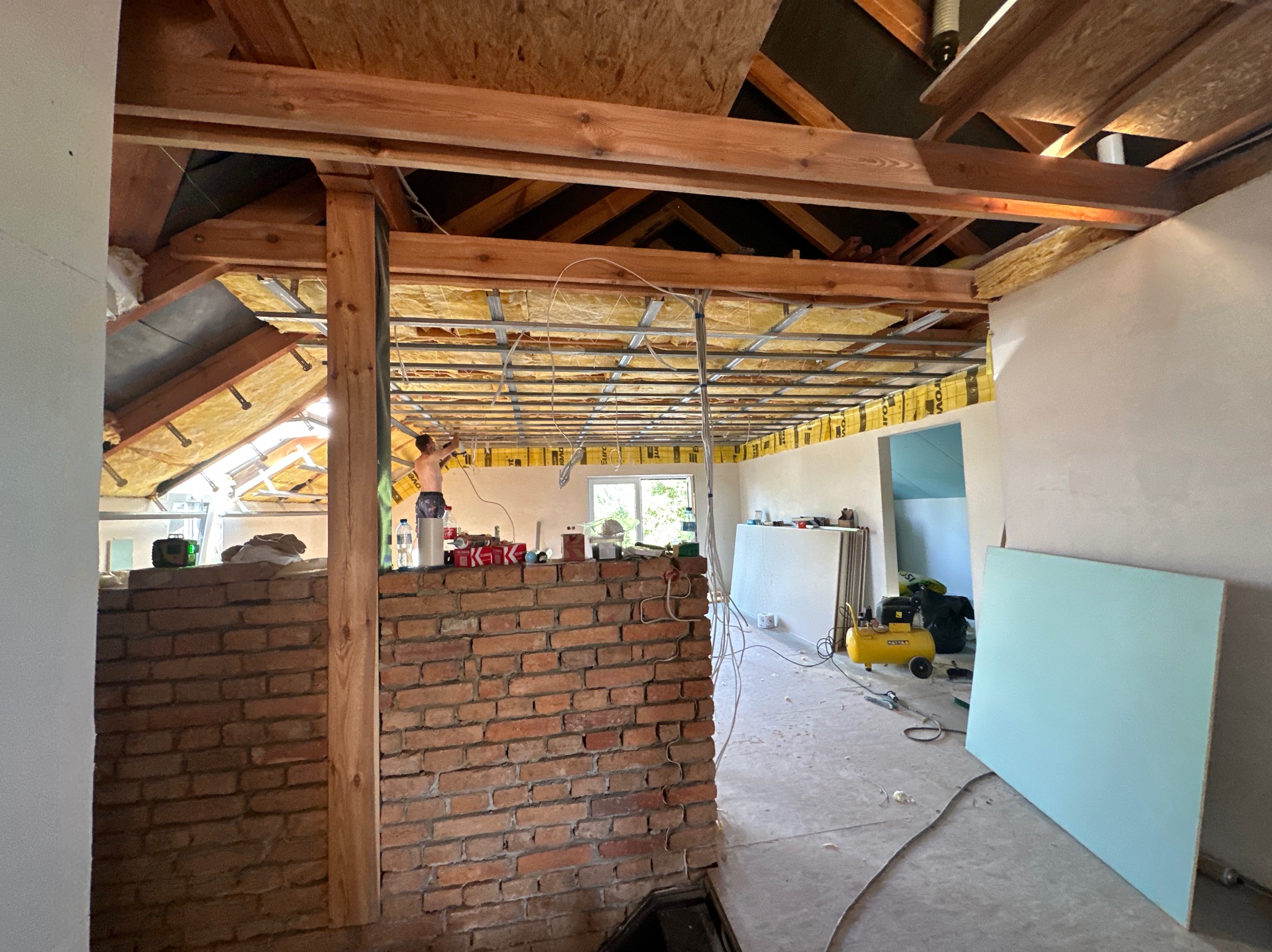 Wnętrze poddasza w trakcie adaptacji: widoczne ocieplenie wełną mineralną, metalowa konstrukcja pod płyty gipsowo-kartonowe, fragment ściany z cegły, kompresor i płyta kartonowo-gipsowa oparta...