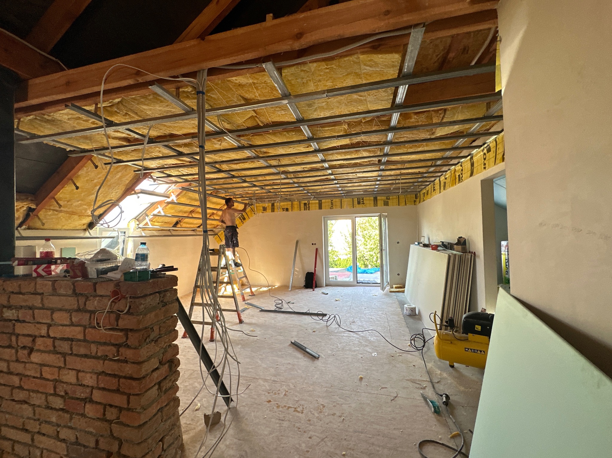 Ocieplanie poddasza wełną mineralną, widoczne metalowe profile konstrukcyjne, pracownik na drabinie montuje izolację, w tle okno dachowe i ściana z oknem balkonowym.