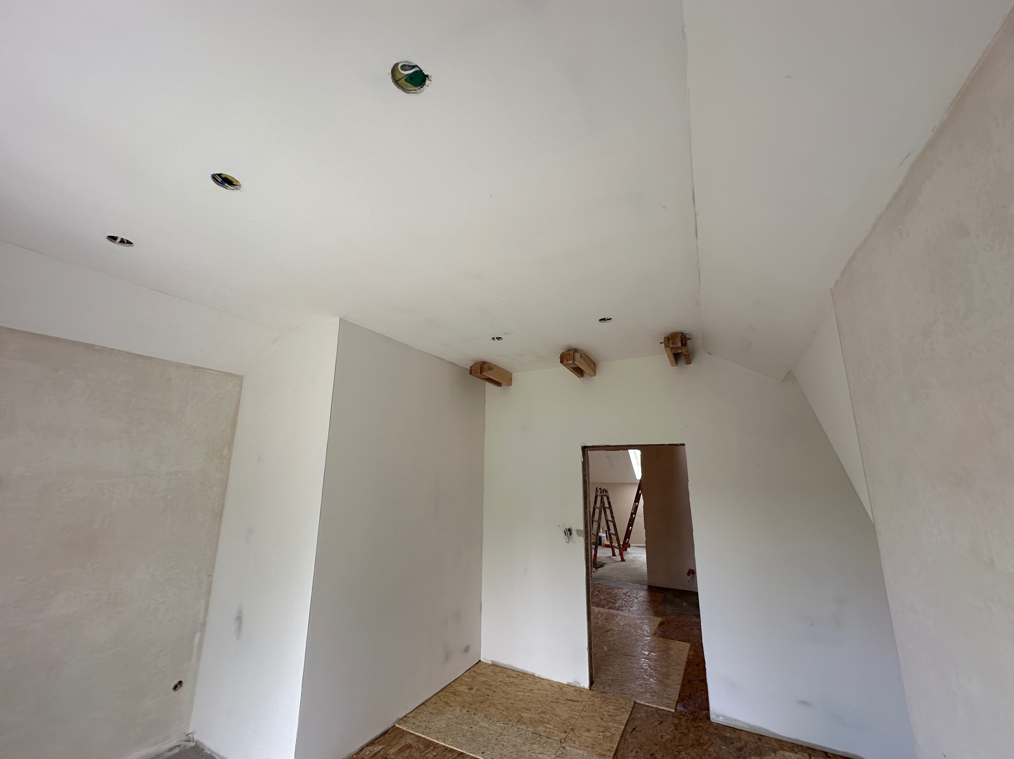 Wnętrze w trakcie prac wykończeniowych: surowe ściany po nałożeniu gładzi, widoczne otwory pod oświetlenie w suficie i drewniane wsporniki, podłoga wyłożona płytami OSB, w tle drabina w przejściu...