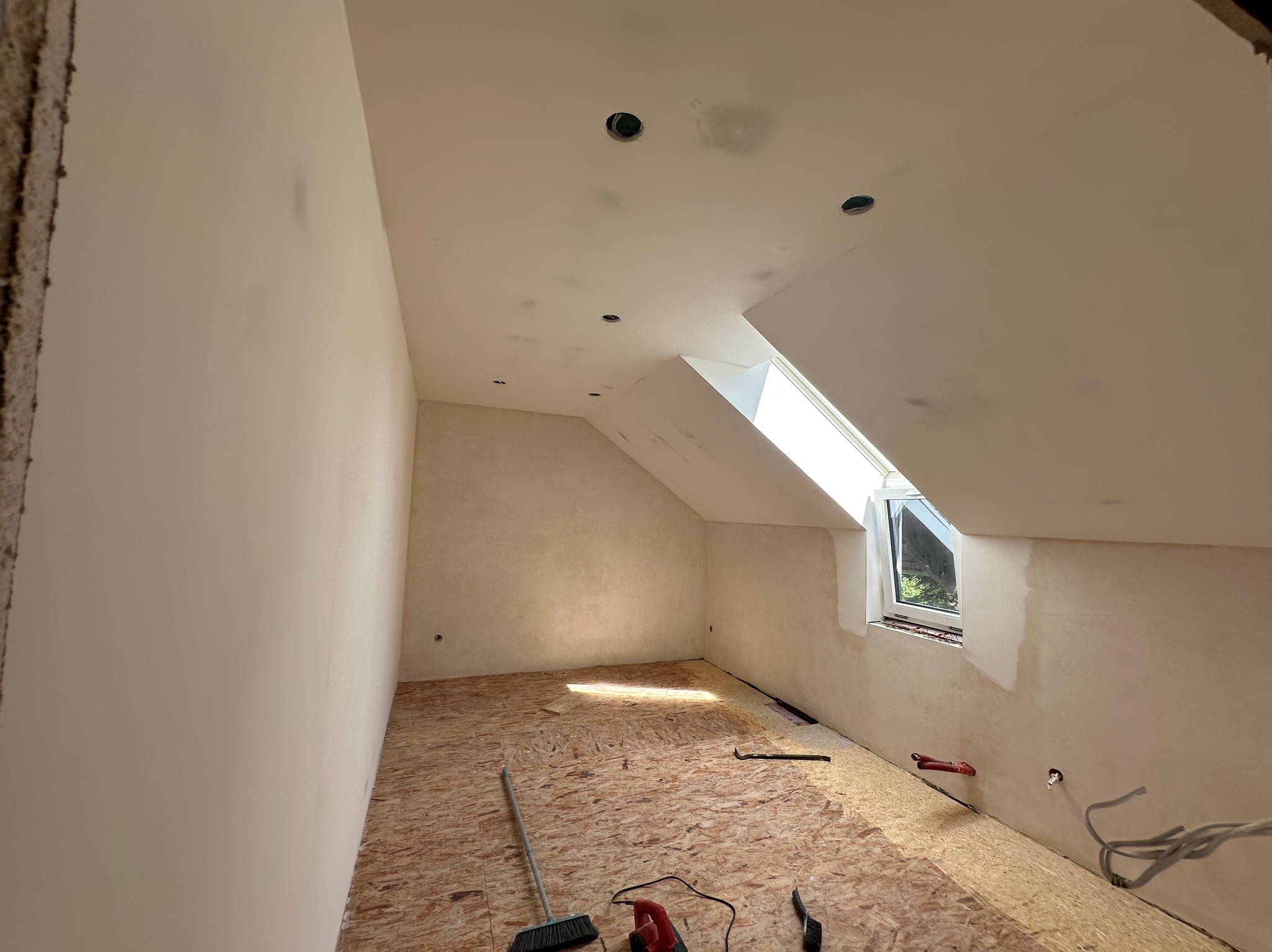 Poddasze w trakcie prac wykończeniowych: ściany po wstępnym wyrównaniu, otwory na oświetlenie w suficie, podłoga wyłożona płytami OSB, widoczne narzędzia i instalacje.