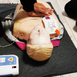 Instruktor demonstruje uciski klatki piersiowej na manekinie podczas szkolenia z pierwszej pomocy, obok leży defibrylator AED z podłączonymi elektrodami.