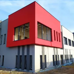 Nowoczesny budynek z jaskrawoczerwoną, kubistyczną nadbudową, elewacja w odcieniach szarości i bieli, teren budowy w nieładzie z widocznymi puszkami farb.