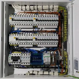 Otwarta skrzynka rozdzielcza z widocznymi bezpiecznikami, przewodami w różnych kolorach (białe, brązowe, niebieskie, żółto-zielone) oraz elementami montażowymi, starannie ułożone wewnątrz białej...