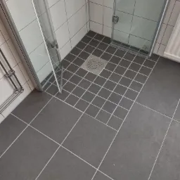 Szare płytki podłogowe w kabinie prysznicowej z odpływem liniowym, białe płytki ścienne w tle.