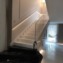 EL-TOM Karol Tomczyk - Eleganckie schody z białego marmuru z podświetleniem LED, szklana balustrada, czarny marmurowy stopień początkowy, kinkiet ścienny w stylu retro, widok na okno z firanką.